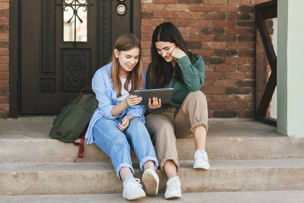 Na schodech u domu sedí dvě krásné dívky-sestry, které přišly z univerzity, používají tablet a smějí se. Technologie, vzdělávání, koncepce volného času — Stock fotografie