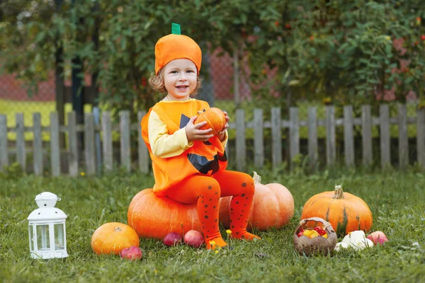 An adorable little girl in Halloween-themed clothing sits on a large orange pumpkin on the lawn and smiles. A girl holds a pumpkin in her hands. Holiday, Halloween. Space for text — Stock Photo, Image