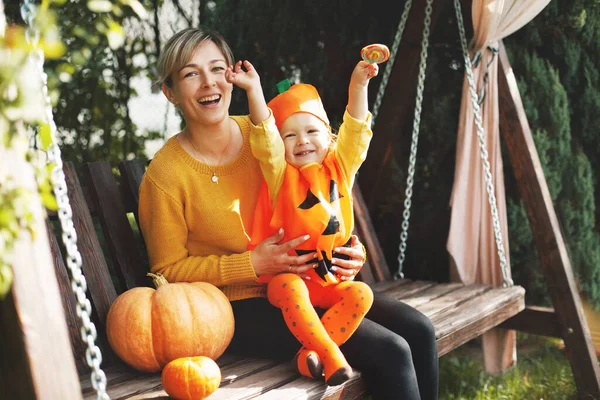 Una giovane madre e la sua figlioletta in costume di Halloween sono sedute su un'altalena in giardino, accanto alle zucche arancioni. Il concetto della vacanza, Halloween — Foto Stock