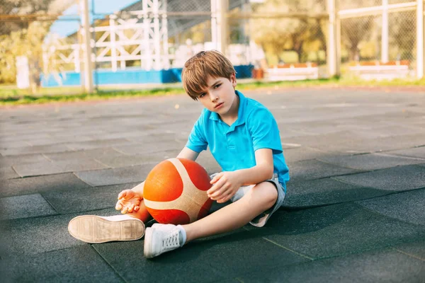 Ein schöner junger Basketballspieler in einem blauen T-Shirt sitzt mit einem Ball in der Hand auf dem Basketballfeld. Der Sportler ruht sich nach dem Training aus. — Stockfoto