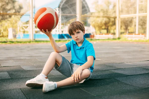 Ein schöner junger Basketballspieler in einem blauen T-Shirt sitzt mit einem Ball in der Hand auf dem Basketballfeld. Der Sportler ruht sich nach dem Training aus. — Stockfoto