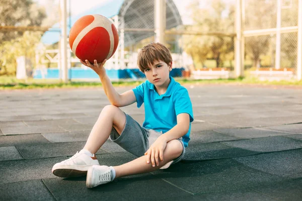 Mavi tişörtlü güzel bir basketbol oyuncusu elinde bir topla basketbol sahasında oturuyor. Sporcu antrenmandan sonra dinleniyor..