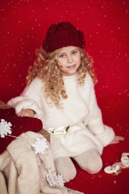 Beyaz kabarık kazaklı, kırmızı şapkalı güzel ve komik kıvırcık bir sarışın büyük bir hediye kutusuyla oturur, kar tanelerini yakalar ve kırmızı bir arka planda gülümser..