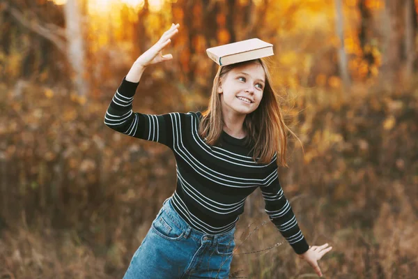 Hermosa rubia se ríe y sostiene un libro sobre su cabeza, caminando en el parque de otoño. — Foto de Stock