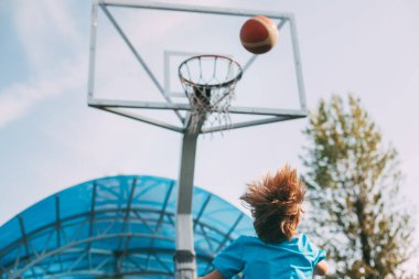 Spor üniformalı bir çocuk basketbol sepetine top atıyor. Bir çocuk basketbol oynuyor. Spor, yaşam tarzı. Arka görünüm, metin için boşluk