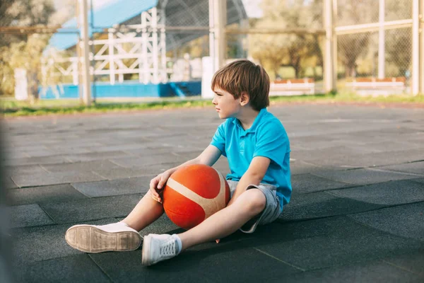 Ein junger Basketballspieler im blauen T-Shirt sitzt mit einem Ball in der Hand auf dem Basketballplatz und ruht sich nach dem Training aus. Das Konzept von Sport und gesundem Lebensstil — Stockfoto