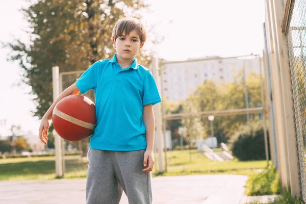Potret seorang anak laki-laki dalam seragam olahraga dengan bola basket di tangannya. Seorang anak laki-laki memegang bola di tangannya setelah bermain basket. Olahraga, pendidikan, gaya hidup sehat — Stok Foto