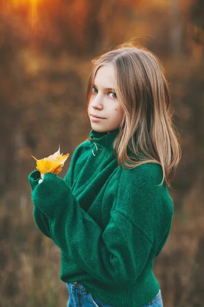Ritratto di una bella adolescente con i capelli biondi e gli occhi azzurri con un sorriso sul viso e una foglia gialla autunnale nel parco — Foto Stock