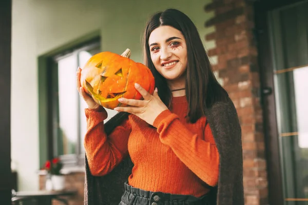 Een mooi meisje met donker haar met make-up voor de viering van Halloween houdt een pompoen in haar handen en glimlacht. — Stockfoto
