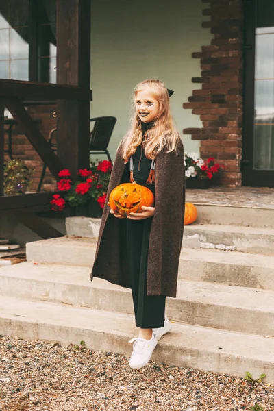 Una piccola ragazza bionda con il trucco per la celebrazione di Halloween tiene in mano una zucca e va a fare una passeggiata per visitare — Foto Stock