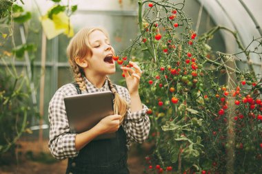 Güzel küçük bir kız elinde bir büyüteçle bir serada duruyor ve sırıtıyor.,