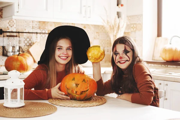 Due ragazze a casa in cucina in costume e trucco per Halloween siedono con una zucca, giocano e ridono. I bambini felici si stanno preparando per Halloween. — Foto Stock