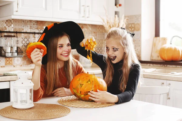 Due ragazze a casa in cucina in costume e trucco per Halloween siedono con una zucca, giocano e ridono. I bambini felici si stanno preparando per Halloween. — Foto Stock