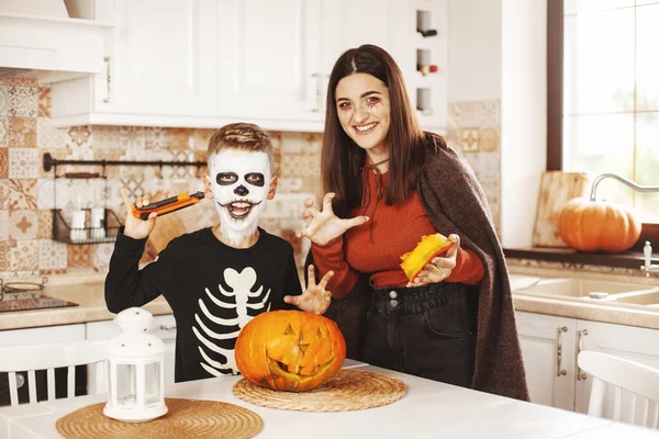 Fratello e sorella felici sono a casa in cucina indossando costumi e trucco di Halloween, intagliando una zucca, giocando e ridendo. — Foto Stock