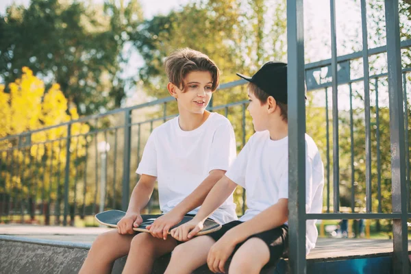 Zwei niedliche Teenager sitzen in einem Skatepark, entspannen sich nach dem Skateboarden und unterhalten sich. Jungen genießen ihre Freizeit im Skatepark, sitzen auf der Rampe. Das Konzept von Jugend, Einheit und Freundschaft — Stockfoto