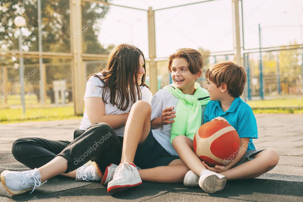 Cheerful high school students sit on the basketball court, relax after the game, talk and laugh. Sports, games, and education. The concept of friendship