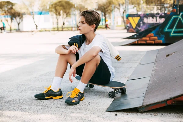 Seorang remaja cantik sedang duduk di skateboard di area khusus taman. Seorang anak laki-laki beristirahat setelah naik di skatepark. Istirahat aktif di udara segar — Stok Foto
