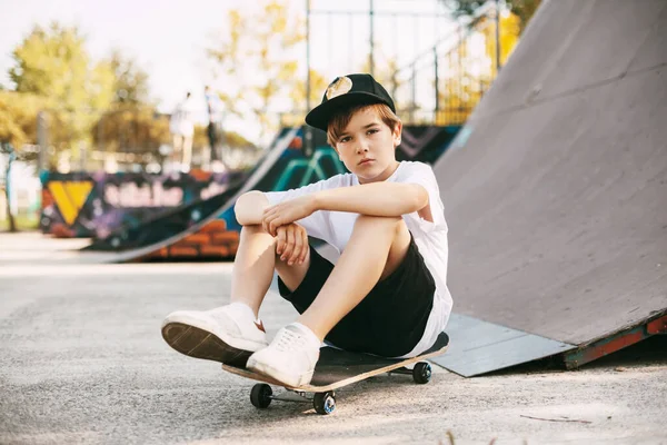 Seorang remaja cantik sedang duduk di skateboard di area khusus taman. Seorang anak laki-laki beristirahat setelah naik di skatepark. Istirahat aktif di udara segar — Stok Foto