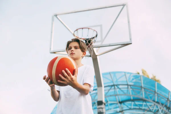 Ένας όμορφος έφηβος με λευκό μπλουζάκι στέκεται στο γήπεδο του μπάσκετ και κρατά ένα μπάσκετ στα χέρια του. Η έννοια του αθλητισμού και ενός υγιεινού τρόπου ζωής — Φωτογραφία Αρχείου