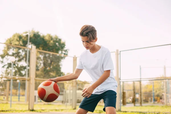 Atlet imut yang fokus memimpin bola dalam permainan basket. Anak laki-laki bermain basket sepulang sekolah. Olahraga, gaya hidup sehat, rekreasi — Stok Foto