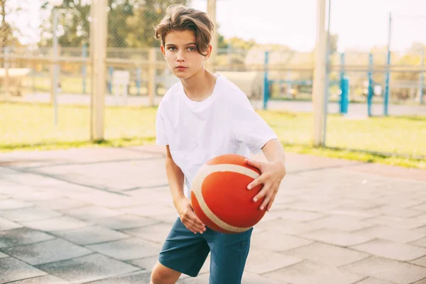एक सफेद टी-शर्ट में एक सुंदर किशोर बास्केटबॉल कोर्ट पर खड़ा है और अपने हाथों में एक बास्केटबॉल रखती है। खेल की अवधारणा और एक स्वस्थ जीवन शैली — स्टॉक फ़ोटो, इमेज
