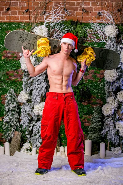 young sexy sporty young man in red pants and Santa's cap keeps snowboard on his shoulders, vertical view