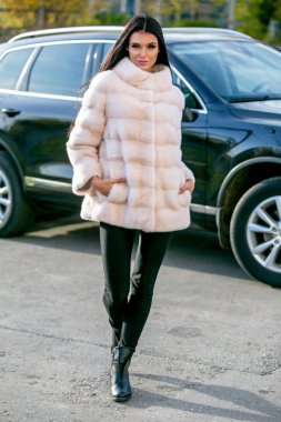 Güzel esmer bir ışık kürk ceket ve siyah pantolon yürüyüş aşağı sokaktaki bir sonbahar güneşli günde araba önünde elleri ona ceplerinde vardır.