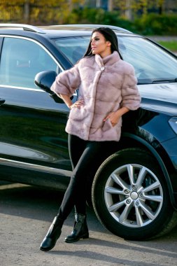 Bir ışık kürk ceket ve siyah pantolon güzel bir esmer bir sonbahar güneşli günde arabanın yakınında duruyor ve gülümseyen, eli cebinde olduğunu