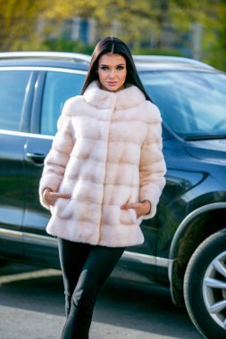 Bir ışık kürk ceket ve siyah pantolon güzel bir esmer bir sonbahar güneşli günde araba önünde sokakta duruyor, ona ceplerinde elleri vardır