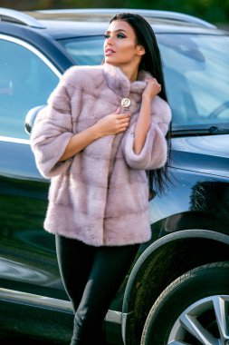 Bir açık renkli kürk ceket ve siyah pantolon güzel bir esmer bir sonbahar güneşli günde bir arabanın yakınında duran, cinsel uzağa bakarak, onu yaka tutarak
