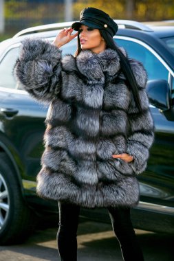 Güzel bir esmer kısa bir ceket, siyah pantolon ve siyah kap arabanın yakınında bir sonbahar güneşli günde duruyor, bir kenara cinsel olarak görünüyor ve poz veriyor