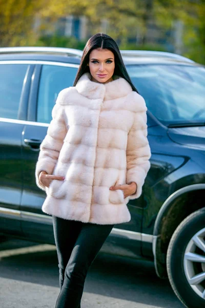 Bir ışık kürk ceket ve siyah pantolon güzel bir esmer bir sonbahar güneşli günde araba önünde sokakta duruyor, ona ceplerinde elleri vardır
