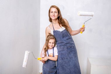 Repair in the apartment. Happy family mother and little daughter in blue aprons paints the wall with white paint. Mom and daughter are holding paint rollers and smiling. Horizontal portrait clipart