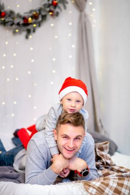 Mutlu aile, baba ve oğlu, yatak odasında sabah Noel için dekore edilmiştir. Babası üzerinde oturup Noel Baba'nın şapkalı çocuk. Yeni yıl ve Noel Tema. Tatil ruh hali.