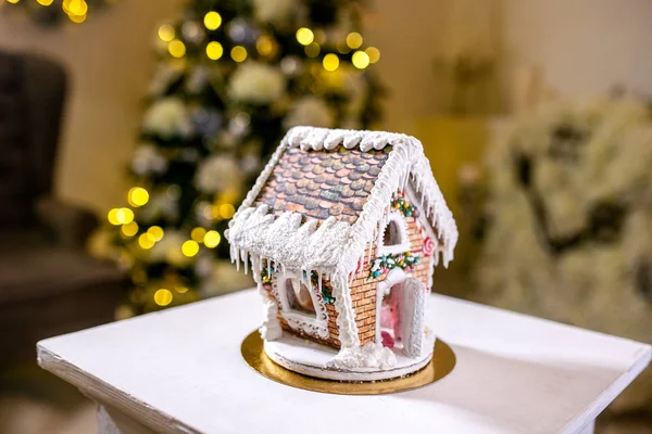 Gingerbread house in front of defocused lights of Christmas decorated fir tree. Holiday sweets. New Year and Christmas theme. Festive mood. Christmas card.