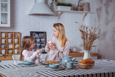 Mutlu küçük kız ve güzel genç annesi kahvaltı birlikte beyaz bir mutfak var. Onlar süt içip kurabiye. Anne bakım ve sevgi. Yatay fotoğraf.