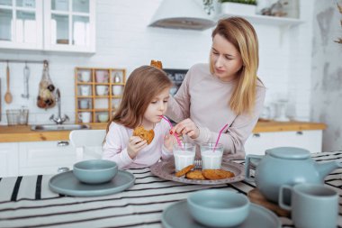 Mutlu küçük kız ve güzel genç annesi kahvaltı birlikte beyaz bir mutfak var. Onlar süt içip kurabiye. Anne bakım ve sevgi. Yatay fotoğraf.