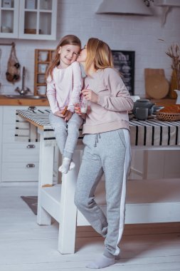 Mutlu küçük kız ve güzel genç annesi kahvaltı birlikte beyaz bir mutfak var. Anne hugs ve kızı öper. Anne bakım ve sevgi. Dikey fotoğraf.