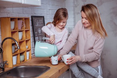 Mutlu küçük kız ve güzel genç annesi kahvaltı birlikte beyaz bir mutfak var. Onlar dökün ve çay iç. Anne bakım ve sevgi. Yatay fotoğraf.