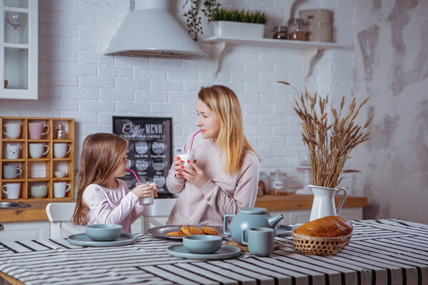 Счастливая маленькая девочка и ее красивая молодая мать завтракают вместе на белой кухне. Они пьют молоко и едят печенье. Материнская забота и любовь. Горизонтальное фото
.