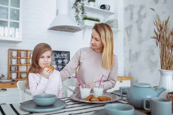 Mutlu küçük kız ve güzel genç annesi kahvaltı birlikte beyaz bir mutfak var. Onlar süt içip kurabiye. Anne bakım ve sevgi. Yatay fotoğraf.