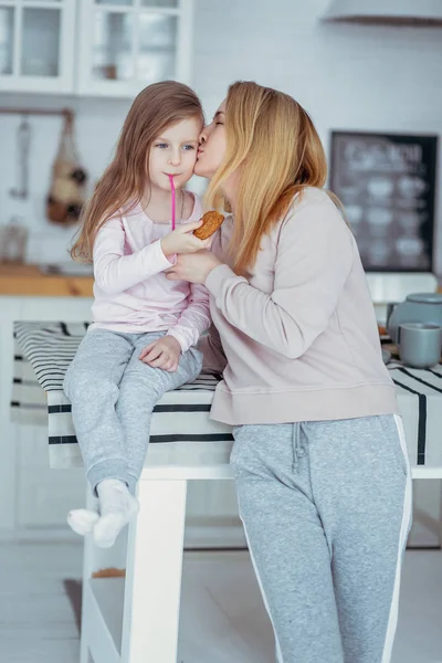 Mutlu küçük kız ve güzel genç annesi kahvaltı birlikte beyaz bir mutfak var. Anne hugs ve kızı öper. Anne bakım ve sevgi. Dikey fotoğraf.