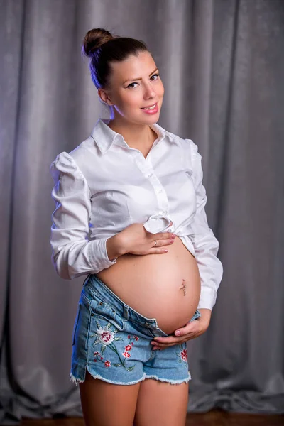 Mulher feliz grávida tocando sua barriga. Retrato de mãe jovem grávida, acariciando a barriga e sorrindo. Conceito de gravidez saudável, fêmea expectante morena em um fundo cinza — Fotografia de Stock