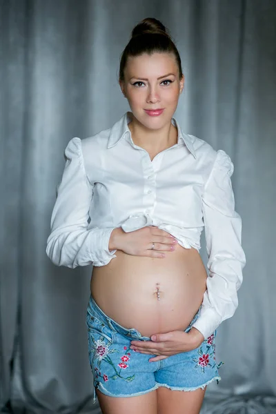 Femme enceinte heureuse touchant son ventre. Portrait de jeune mère enceinte, caressant son ventre et souriant. Concept de grossesse saine, femme enceinte brune sur fond gris — Photo
