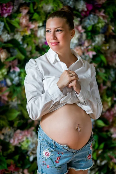 Gravid glad kvinna röra hennes mage. Gravid ung mamma porträtt, smekande hennes mage och leende. Frisk graviditet koncept, brunett gravid kvinna på blommor bakgrund — Stockfoto