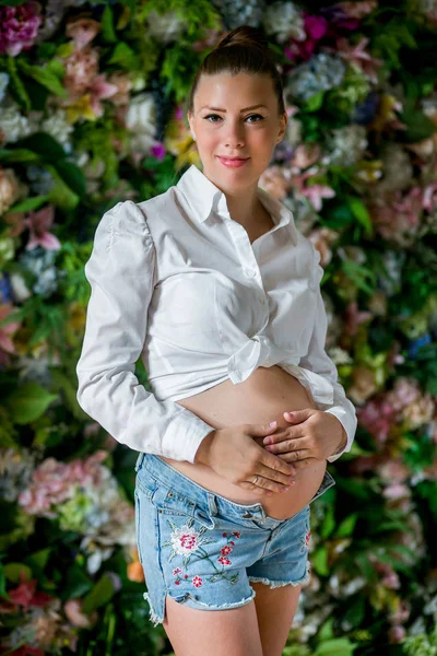 Gravid glad kvinna röra hennes mage. Gravid ung mamma porträtt, smekande hennes mage och leende. Frisk graviditet koncept, brunett gravid kvinna på blommor bakgrund — Stockfoto