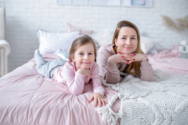Güzel genç anne ve küçük kızı yatak odasında yatağın üstünde yan yana yatarken, oynarken, sarılma ve eğleniyor. Anne bakım ve sevgi. Yatay fotoğraf