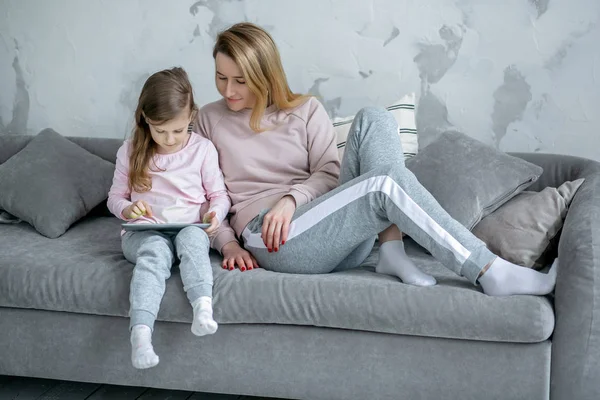 Beautiful young mother and her little daughter are watching movies together and playing on the tablet while sitting on the sofa. Mom hugs daughter. Maternal care and love. Horizontal photo