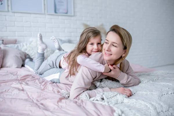 Hermosa madre joven y su hija pequeña están acostados juntos en la cama en el dormitorio, jugando, abrazándose y divirtiéndose. Cuidado materno y amor. Fotografía horizontal — Foto de Stock