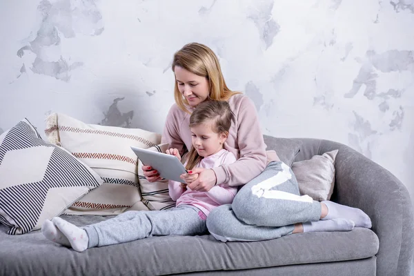 Vacker ung mamma och hennes lilla dotter titta på filmer tillsammans och spela på tabletten medan du sitter i soffan. Mamma kramar dotter. Mödravård och kärlek. Horisontella Foto — Stockfoto